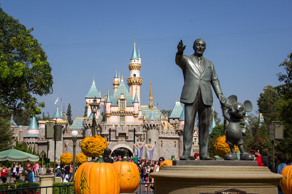 A statue of Walt Disney holding Mickey Mouse's in front of the Disneyland castle.